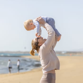 Mother enjoying summer vacations holding, playing and lifting his infant baby boy son high in the air on sandy beach on Lanzarote island, Spain. Family travel and vacations concept