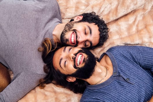 top view of a happy gay male couple laughing lying on the bed with their faces together, concept of real people lifestyle at home and love between people of the same sex