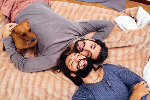 happy gay male couple smiling lying with their little dog on the bed with faces together, concept of real people lifestyle at home and love between people of the same sex