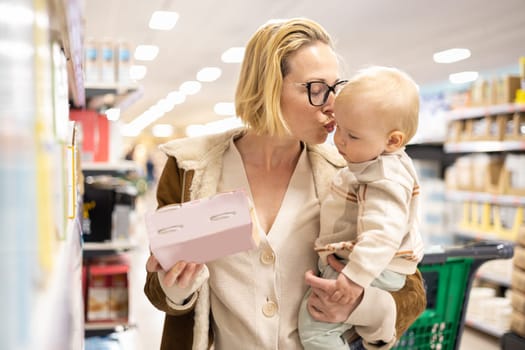 Caucasian mother shopping with her infant baby boy child choosing products in department of supermarket grocery store