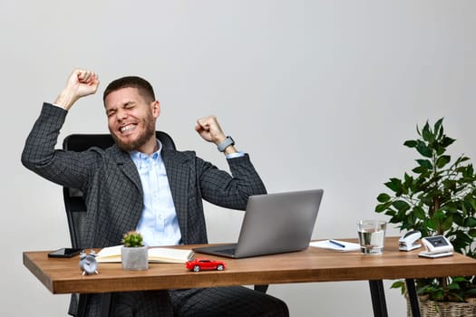 Satisfied young man gesturing emotionally and celebrating victory while sitting on chair at desk, using laptop pc computer