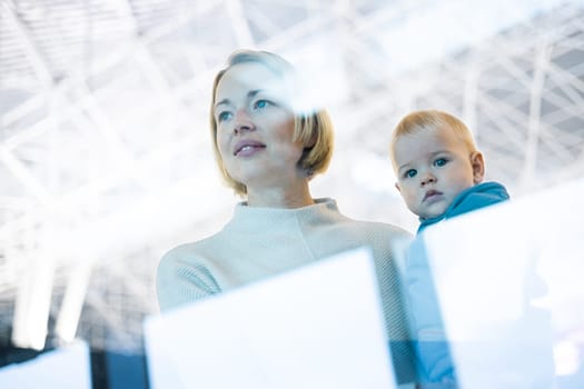 Thoughtful young mother looking trough window holding his infant baby boy child while waiting to board an airplane at airport terminal departure gates. Travel with baby concept