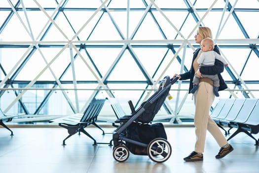 Mother carying his infant baby boy child, pushing stroller at airport departure terminal moving to boarding gates to board an airplane. Family travel with baby concept