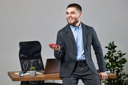 Successful caucasian cheerful man holding a toy car while standing at desk. Car dealerships offer car . Car insurance