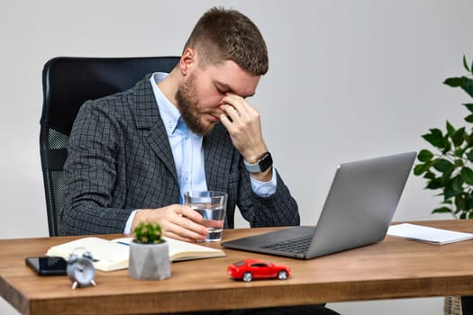 bearded man sitting on chair at desk and suffering from headache at workplace, using laptop