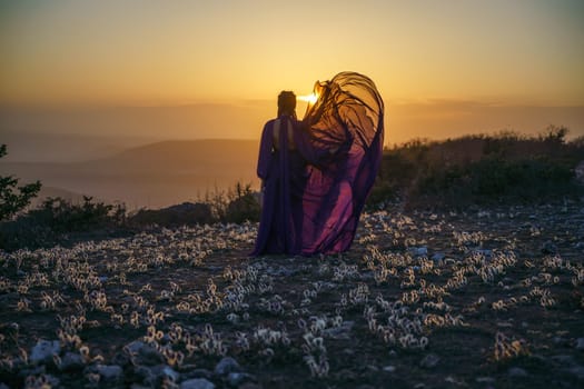 Sunset purple dress woman mountains. Rise of the mystic. sunset over the clouds with a girl in a long purple dress. In the meadow there is a grass dream with purple flowers