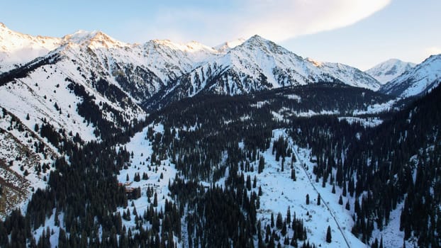 Coniferous forest in snowy mountains. White clouds float across the sky. Top view from a drone. In places, the road to the mountains and buildings are visible. There is a pipe along the gorge. Sunset