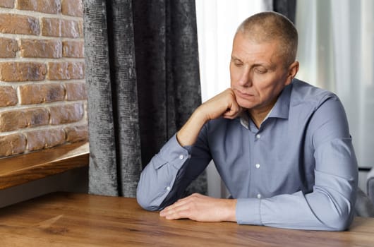 Portrait of a pensive middle-aged man sitting at a table. The man leaned his head on his hand looking down.
