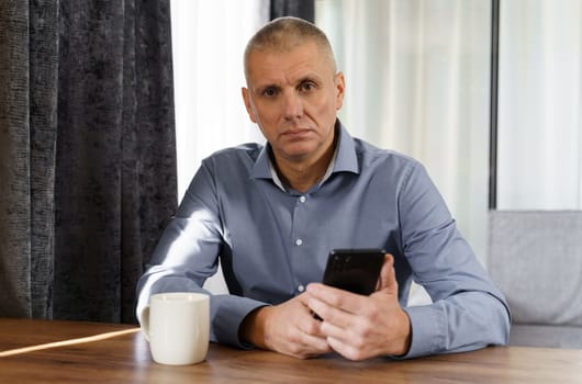 Portrait of a male businessman who sits at a table, holds a phone in his hands, looks into the camera.