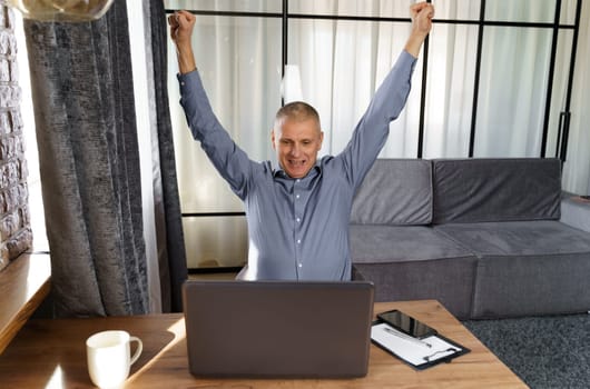 A businessman working at a computer rejoices in a perfect successful transaction, raising his hands up. The concept of success.