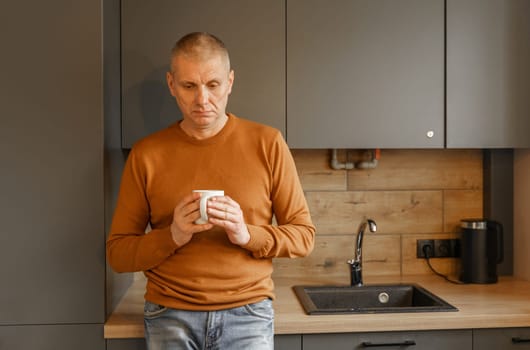 Portrait of a mature man in an orange jumper in the kitchen with a mug of warm tea.