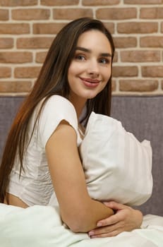 Young woman hugging a pillow on a comfortable bed with white linens. Vertical frame.