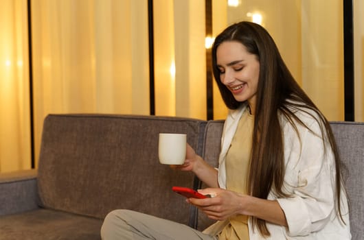 Young woman chatting with friends, surfing on the phone, enjoying her weekend at home with a cup of tea.