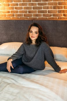 An attractive woman is sitting on a bed in a bedroom wearing blue trousers and a gray jumper. Smiling and looking at the camera. Vertical frame.