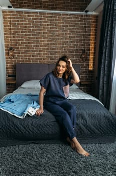 A beautiful woman in designer clothes sits on a bed, clothes lie nearby. Vertical frame.