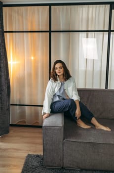 Portrait of a beautiful young woman who is sitting on the back of the sofa, looking at the camera and smiling. Vertical frame.