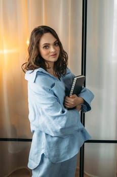 A portrait of a young woman in a light blue suit is standing in a room with a tablet and a notebook in her hands. Looks into the camera. Vertical frame.