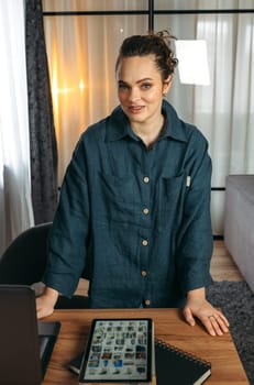 Portrait of a business woman standing near the desk. Notepad, tablet and laptop on the table. Vertical frame.