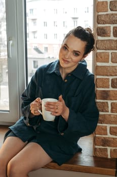 Portrait of a beautiful woman who is sitting on the windowsill indoors with a white cup in her hands. Vertical frame.