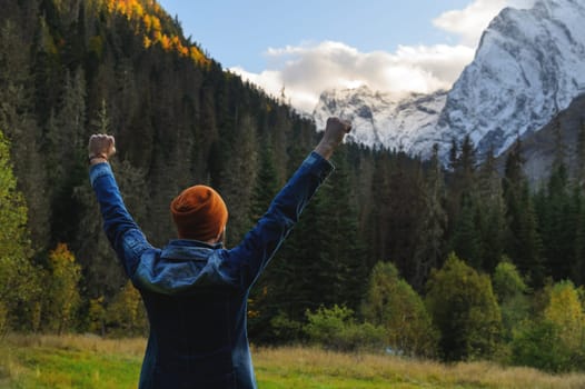 A man stands against the backdrop of a mountain landscape with his hands raised high. Autumn or summer, in the foreground a dense green forest, behind it are snow-capped mountains, a joyful tourist.