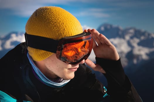 Portrait of a professional skier athlete in a blue jacket, black and orange mask looks to the side. Background with snow covered mountains of ski resort.