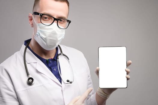A young and handsome smart doctor wearing a surgical hygiene protective mask on a gray background and holding a tablet computer in his hand with a blank mockup screen. Idea for doctors.