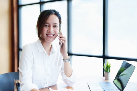 Portrait of a young Asian woman showing a smiling face as she uses her phone, computer and financial documents on her desk in the early morning hours.