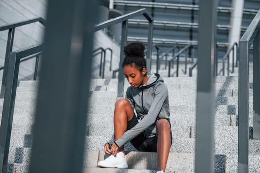 Sits on stairs. Young african american woman in sportive clothes have workout outdoors at daytime.