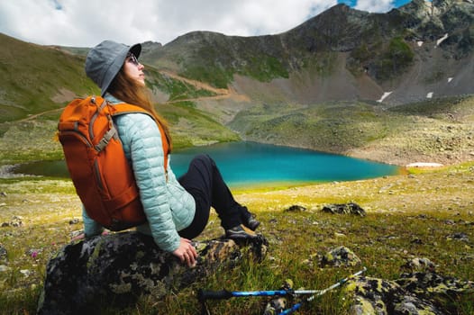 A woman sits in a vantage point above a stunning turquoise lake surrounded by mountains during the day. She sits on a boulder on the side with a backpack while relaxing on a hike.
