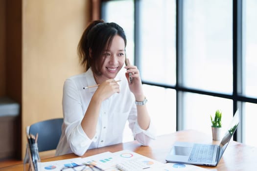 Portrait of a young Asian woman showing a smiling face as she uses her phone, computer and financial documents on her desk in the early morning hours.