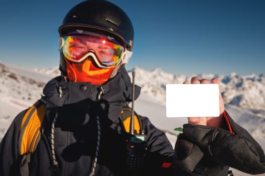 A snowboarder holds an empty lift pass with a mountain in the background. Blank ski pass in the hand of a young skier in winter gear looking at the camera. Concept illustrating ski entry fee.