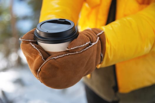 A woman in gloves holds a paper cup of coffee in a winter city in a park outdoors. Hot and warming drinks in winter concept. hands close up.