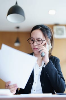 Portrait of a young Asian woman showing a serious face as she uses her phone, financial documents and computer laptop on her desk in the early morning hours.