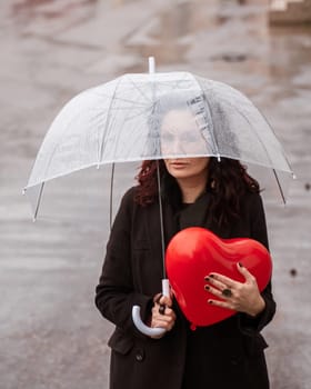 Sad middle-aged woman in a black coat and boots under a transparent umbrella. He holds a balloon in the form of a heart in his hands. Rain in the city, outdoor scene, water drops on the umbrella. The concept of loneliness, unhappy love
