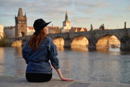 Stylish young beautiful woman earing black hat in Prague with Charles Bridge on background. Elegant retro lady fine art portrait.