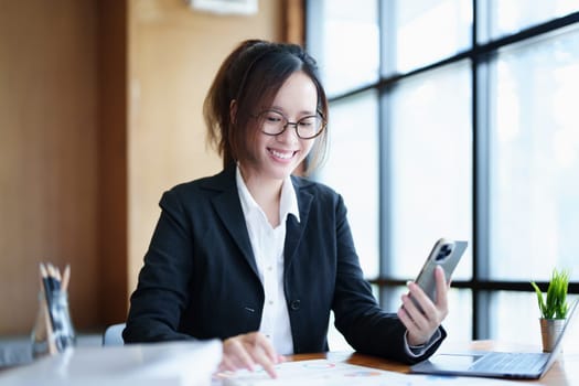 Portrait of a young Asian woman showing a smiling face as she uses her phone, computer and financial documents on her desk in the early morning hours.