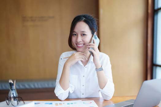 Portrait of a young Asian woman showing a smiling face as she uses her phone, computer and financial documents on her desk in the early morning hours.