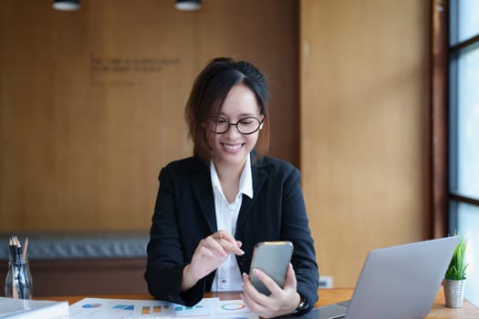 Portrait of a young Asian woman showing a smiling face as she uses her phone, computer and financial documents on her desk in the early morning hours.