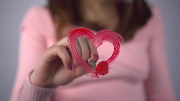 A young woman draws a heart on the glass. A girl in a pink sweater draws with a brush and red paint. Close-up. 4k