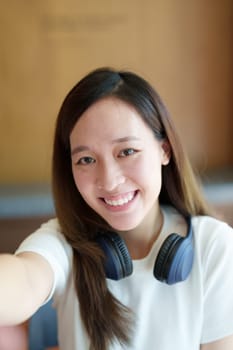 A portrait of a young Asian woman wearing headphones showing a smiling face taking a selfie.