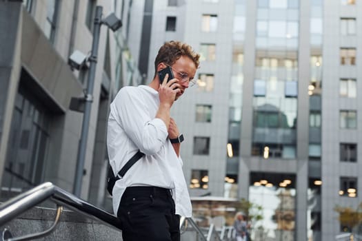 Have conversation by the phone. Young elegant man in good clothes is outdoors in the city at daytime.