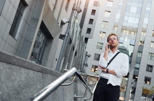 Have conversation by the phone. Young elegant man in good clothes is outdoors in the city at daytime.