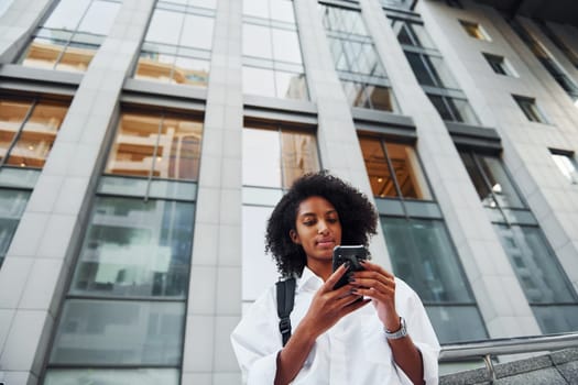 Uses phone. African american woman in good clothes is outdoors in the city at daytime.