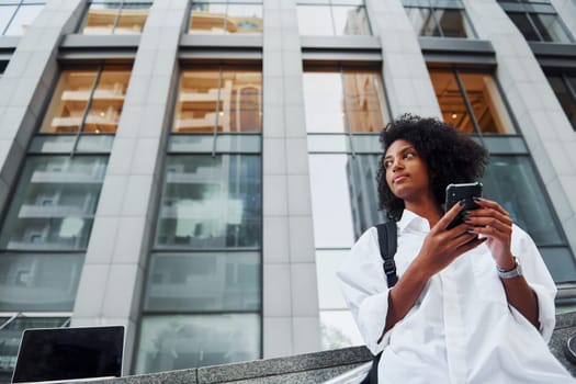 Uses phone. African american woman in good clothes is outdoors in the city at daytime.