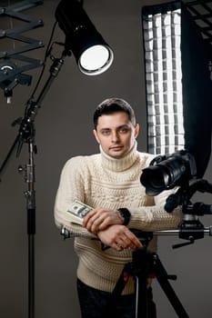 Professional handsome photographer with digital camera on tripod holding hundred dollar bills on gray background with lighting equipment