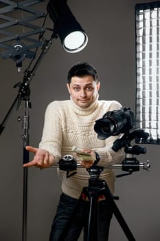 surprised professional photographer with digital camera on tripod holding hundred dollar bills on gray background with lighting equipment