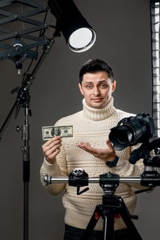 Professional handsome photographer with digital camera on tripod showing money on gray background with lighting equipment