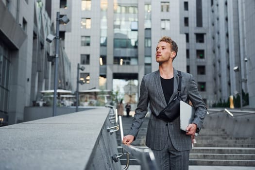 Laptop in hands. Young elegant man in good clothes is outdoors in the city at daytime.