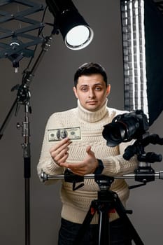 Professional handsome photographer with digital camera on tripod showing hundred dollar bill on gray background with lighting equipment