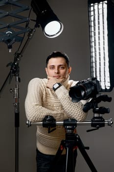 Positive professional photographer with digital camera on tripod on gray background with lighting equipment